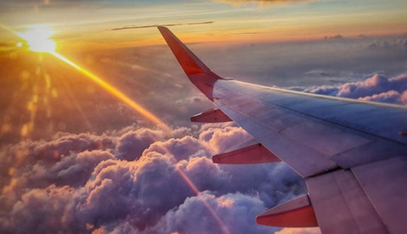 View from airplane window; wing visible against a vibrant sunset sky. Golden sun rays illuminate fluffy pink clouds, evoking a serene, dreamy mood.